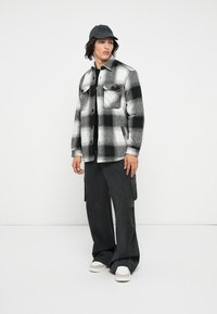 Cămașă flannel cu carouri alb-negru, cu două buzunare pe piept, asortată cu pantaloni cargo negri și adidași albi; model purtând o șapcă neagră.