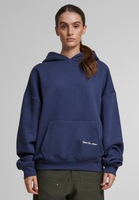 Marineblauwe hoodie van zacht materiaal met een kangoeroezak, oversized pasvorm en ribgebreide manchetten; voorzien van een wit geborduurd merklogo.