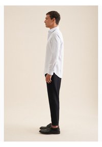 Witte lange mouwen shirt met een knoopsluiting kraag, gecombineerd met marineblauwe tapered broek en zwarte schoenen, bekeken vanaf de zijkant.