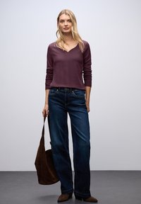 Donna che indossa una maglietta lunga viola, jeans blu scuro e stivali marroni, mentre tiene un grande borsa in suede marrone al suo fianco.