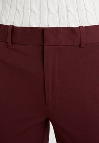 Gros plan sur un pantalon sur mesure bordeaux avec passants de ceinture porté sur un pull blanc en maille torsadée.