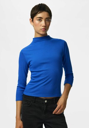 Ung kvinde med kort mørkt hår iført en knaldblå top med trekvartærmer og sorte jeans mod en ensfarvet lys baggrund.