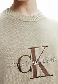 Pull beige avec col côtelé, arborant le logo brodé marron "CK CALVIN KLEIN JEANS" sur le devant dans un design contrastant.