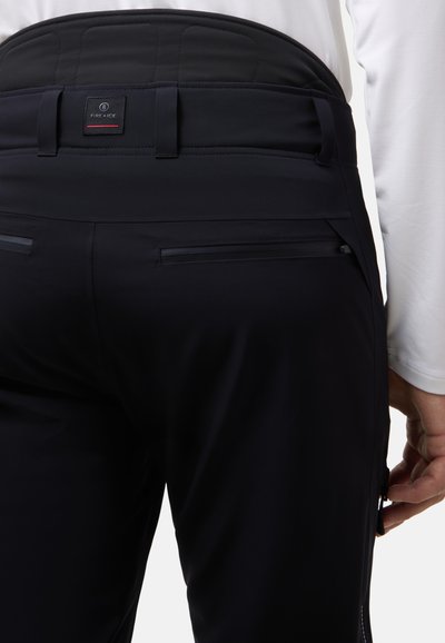 Detail osoby v černých kalhotách se zipovými zadními kapsami a vycpaným páskem s nápisem "B FIRE+ICE", která si upravuje boční zip.