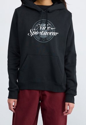 Osoba ubrana w czarną bluzę z kapturem Nike Sportswear z przednią kieszenią i bordowe spodnie, widoczna od szyi do bioder na jednolitym tle.