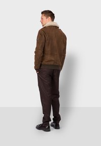 Giacca in suede marrone con colletto in shearling, dettagli con tasche laterali e polsini a coste. Abbinata a pantaloni scuri e scarpe nere.