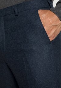 Ruka spočívá uvnitř přední kapsy tmavě námořnických vlněných kalhot s viditelným pasem a šedým úpletem na rukávu.