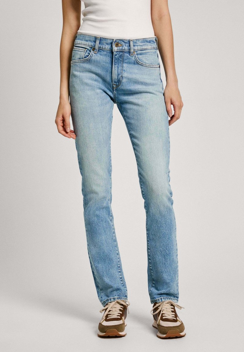 Light blue jeans made of denim, featuring a fitted shape, five pockets, and slight fading. Paired with brown and white sneakers.