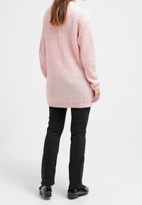 Pull en tricot rose à manches longues et coupe ample avec un motif texturé, associé à un pantalon noir et des chaussures plates noires.