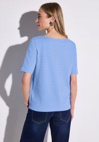 Blå texturerad topp med korta ärmar, rund halsringning och en något lös passform, matchad med mörk denimjeans. Inga synliga mönster eller detaljer.