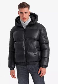 Zwarte pufferjack met een capuchon, glanzende textuur, voorkant met ritssluiting, elastische manchetten en zijzakken. Draagt over een grijze fleece top.