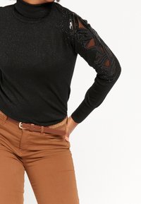 Svart polotröja med glittrande textur och genomskinliga ärmar med utskärningar som har svarta detaljer. Matchad med bruna byxor och ett flätat bälte.