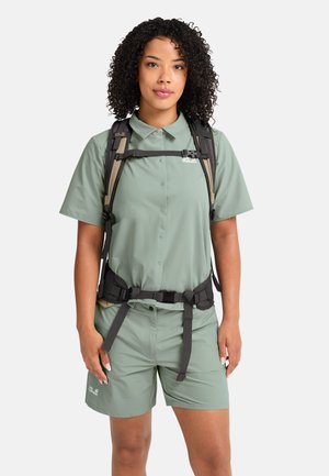 Vrouw met krullend haar, gekleed in een saliegroene wandelshirt en korte broek, uitgerust met een zwart-beige rugzakriem tegen een witte achtergrond.