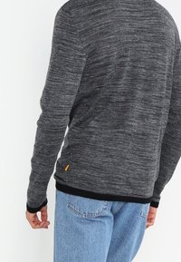 Gråmelerad tröja med långa ärmar, rund halsringning och svart ribbad nederkant. En liten orange logotyp syns nära nederkantens vänstra sida.