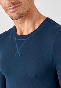 Maglietta a maniche lunghe blu navy realizzata in tessuto morbido, con scollatura tonda e cuciture blu a contrasto disposte a "X" vicino al petto.
