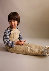 Salopette en velours côtelé beige associée à une chemise à rayures grises et blanches à manches longues, un enfant assis avec des cheveux bruns courts et des chaussures blanches.