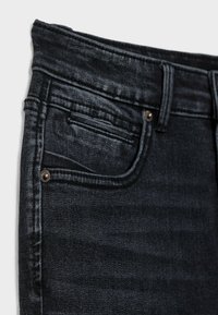 Jean en denim noir avec une petite poche avant, tissu texturé, coutures visibles et détails en bronze sur la ceinture.