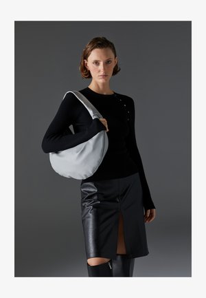 Hobo-veske i lys grå, mykt materiale, rund form. Stylet med en svart langermet topp og et svart skinnskjørt med en frontsplitt.