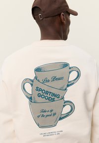 Κρεμ φούτερ με γραφικό στοιχείο με επάλληλα φλιτζάνια καφέ σε μπλε χρώμα και κείμενο «Les Deux», «SPORTING GOODS» και «Πιες μια γουλιά από την καλή ζωή».