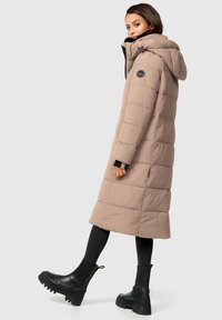 Abrigo largo acolchado en marrón claro, con capucha y diseño acolchado. Combinado con botas negras hasta el tobillo y pantalones negros ajustados.