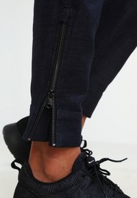 Granatowe spodnie dresowe z podwójnymi zameczkami po bokach na mankietach, wykonane z teksturowanej tkaniny; zestawione z czarnymi butami sportowymi z subtelnym wzorem.