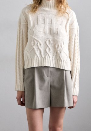 Personne portant un pull à col roulé en maille torsadée crème et un short taupe taille haute sur un fond uni.