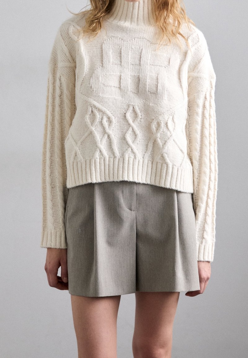 Personne portant un pull à col roulé en maille torsadée crème et un short taupe taille haute sur un fond uni.