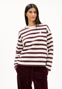 Junge Frau mit langen schwarzen Haaren, die ein cremefarbenes und kastanienbraunes gestreiftes Langarmshirt und kastanienbraune Cordhosen trägt, mit den Händen in den Taschen.