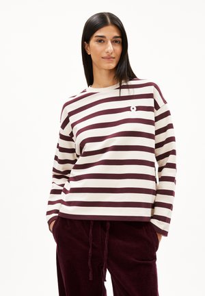 Jonge vrouw met lang zwart haar, gekleed in een crèmekleurige en kastanjebruine gestreepte longsleeve en kastanjebruine corduroybroek, met haar handen in haar zakken.