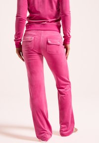 Vibrant roze velours trainingspak met een aansluitend bovenstuk en uitlopende broekspijpen. De broek heeft twee achterzakken met drukknoopsluitingen. Gladde textuur.