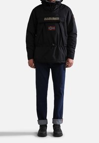 Homme portant une veste à capuche noire avec un patch du drapeau norvégien, un jean bleu foncé retroussé et des chaussures de randonnée noires, debout devant un fond blanc.