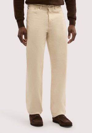 Ein Mann trägt beigefarbene gerade geschnittene Hosen, dunkelbraune Loafers und ein braunes Langarmoberteil und steht vor einem einfarbigen Hintergrund.