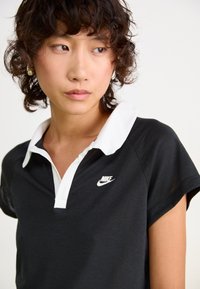 Čierna polo košeľa s bielym golierom a krátkymi rukávmi. Obsahuje malé biele logo Nike na hrudi. Textúrovaný bavlnený materiál.