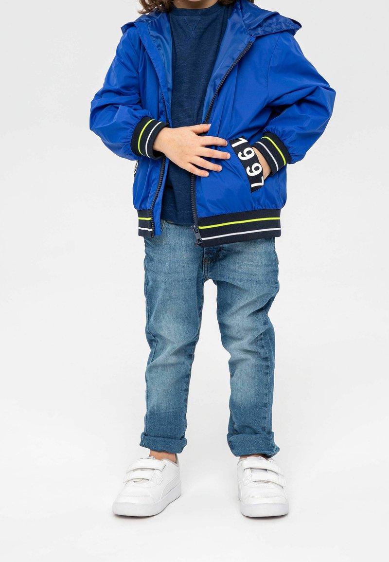 Enfant en veste bleue avec "1991" sur la manche, chemise bleu foncé, jean retroussé et baskets blanches à velcro debout sur fond blanc.