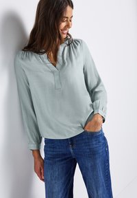 Blouse vert clair à manches longues avec un col en V, dotée d'un tissu texturé et de détails froncés aux épaules, associée à un jean bleu.