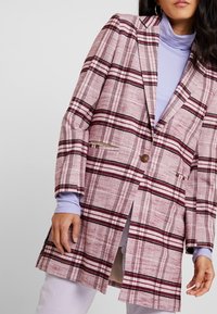 Kariertes Blazer in pinkem Stoff mit kastanienbraunen und weißen Streifen, ein Knopfverschluss und zwei Seitentaschen. Trägt man über einem helllila Oberteil.