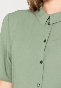 Kortärmad skjorta i ljusgrön tyg har en knapp-up framtill med svarta knappar och en spetsig krage med en texturerad finish.
