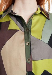 Kariertes Button-up-Hemd in geometrischem Muster in Grüntönen, Braun und Schwarz mit glänzender Oberfläche und goldfarbigen Knöpfen.