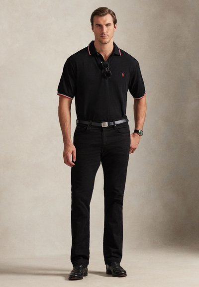 Férfi áll, fekete galléros pólót visel piros-fehér szegéllyel, fekete farmert, fekete övet, fekete cipőt, az inget díszítő napszemüveget és karórát.