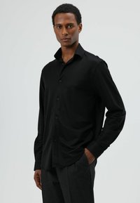 Chemise noire à manches longues en tissu lisse, avec patte de boutonnage, col pointu et coupe décontractée, associée à un pantalon gris texturé.