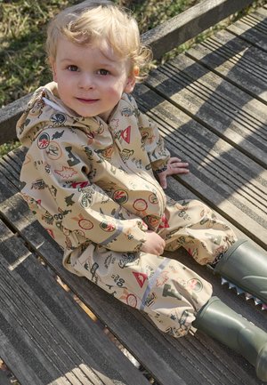 Tout-petit aux cheveux blonds bouclés portant une combinaison de pluie beige à motifs et des bottes vertes, assis sur un banc en bois à l'extérieur sous le soleil.