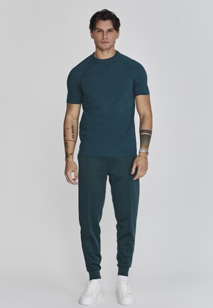 Türkise Sport-T-Shirt mit kurzen Ärmeln, kombiniert mit passenden türkisen Jogginghosen. Das Modell trägt weiße Sneakers und zeigt sichtbare Tattoos.