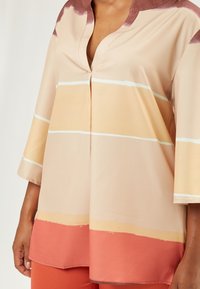 Top ligero, beige, con escote en pico, mangas de tres cuartos y rayas horizontales en suaves tonos de naranja y melocotón. Textura suave.
