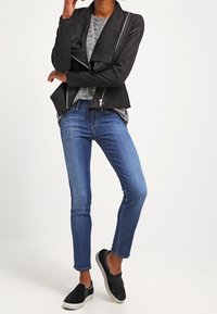 Chaqueta negra corta con un cuello caído y cremalleras diagonales, combinada con jeans ajustados azules y zapatos negros sin cordones, sobre un fondo neutro.