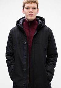 Jonge man in een zwart hoodie-jack over een bordeauxrode quarter-zip trui, staande tegen een effen witte achtergrond.