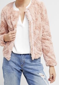 Różowa kurtka z sztucznego futra o prążkowanej fakturze, okrągłym dekolcie, zapięciu na zatrzask i długimi rękawami, zestawiona z białą bluzką i przetartymi dżinsami.