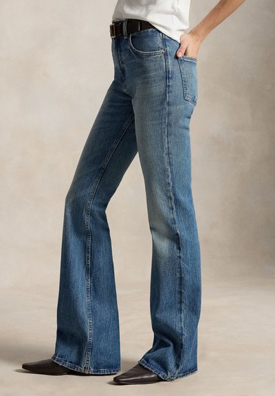 Persona con vaqueros acampanados azules con cinturón negro y zapatos marrones puntiagudos, de pie con una mano en el bolsillo.