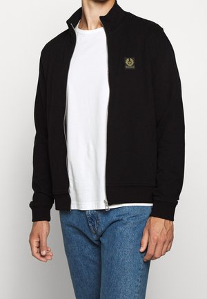 Man draagt een zwarte jack met rits over een wit T-shirt en blauwe spijkerbroek, staand tegen een effen lichte achtergrond.