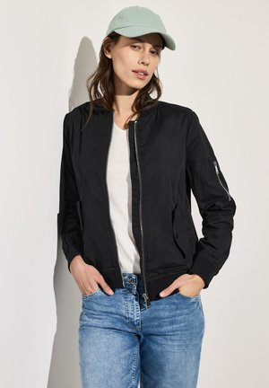 Schwarze Bomberjacke mit Reißverschluss, seitlichen Taschen und elastischen Bündchen. Getragen über einem weißen Shirt und kombiniert mit hellblauen Jeans.