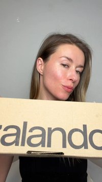 Vrouw met lang haar houdt een grote kartonnen doos vast met gedeeltelijk zichtbare merknaam "zalando" tegen een effen grijze achtergrond.